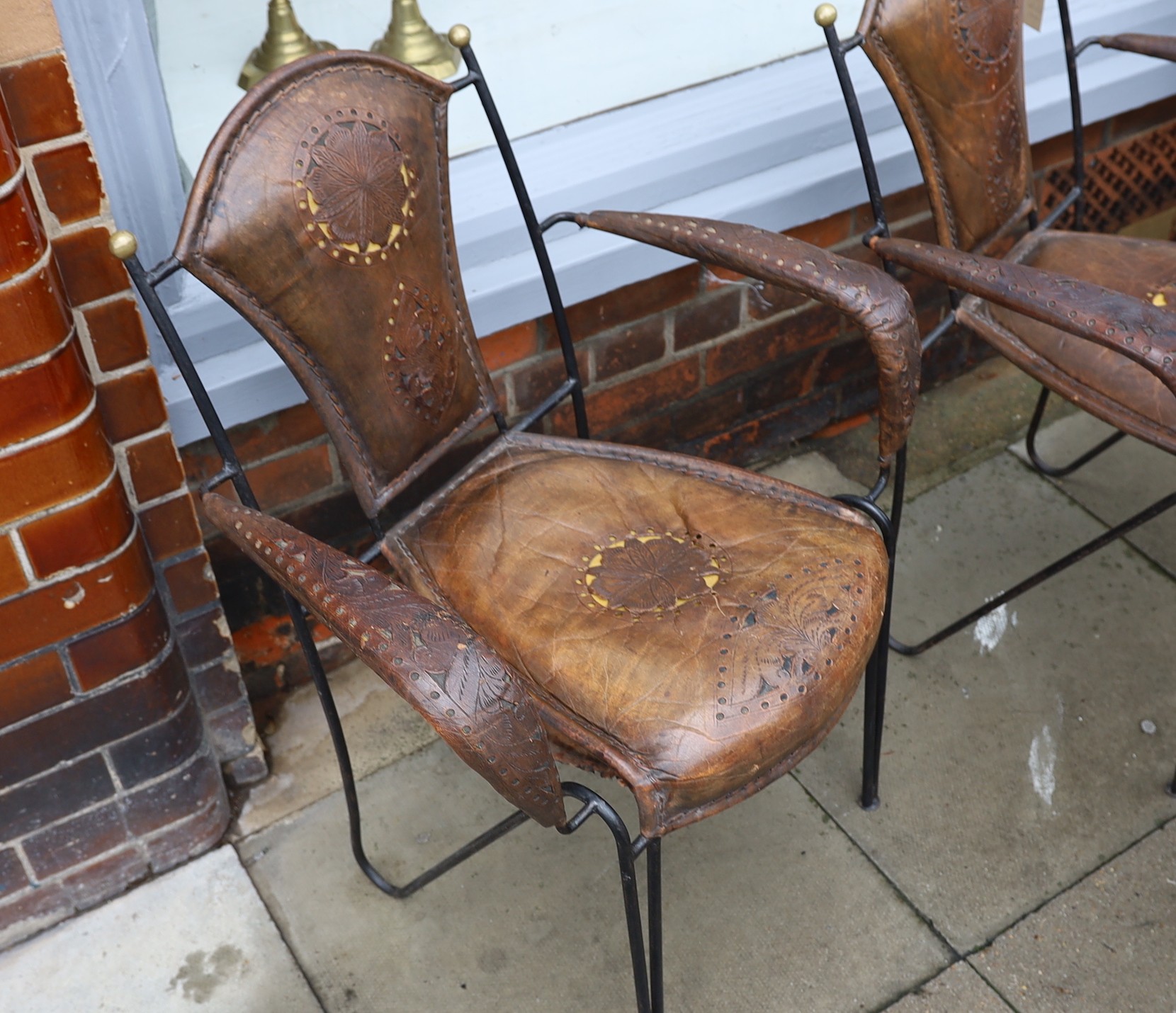 A set of six French wrought iron and brown leather elbow chairs, with brass ball finials, four with tooled and pierced decoration the others plain, height 84cm. width 61cm